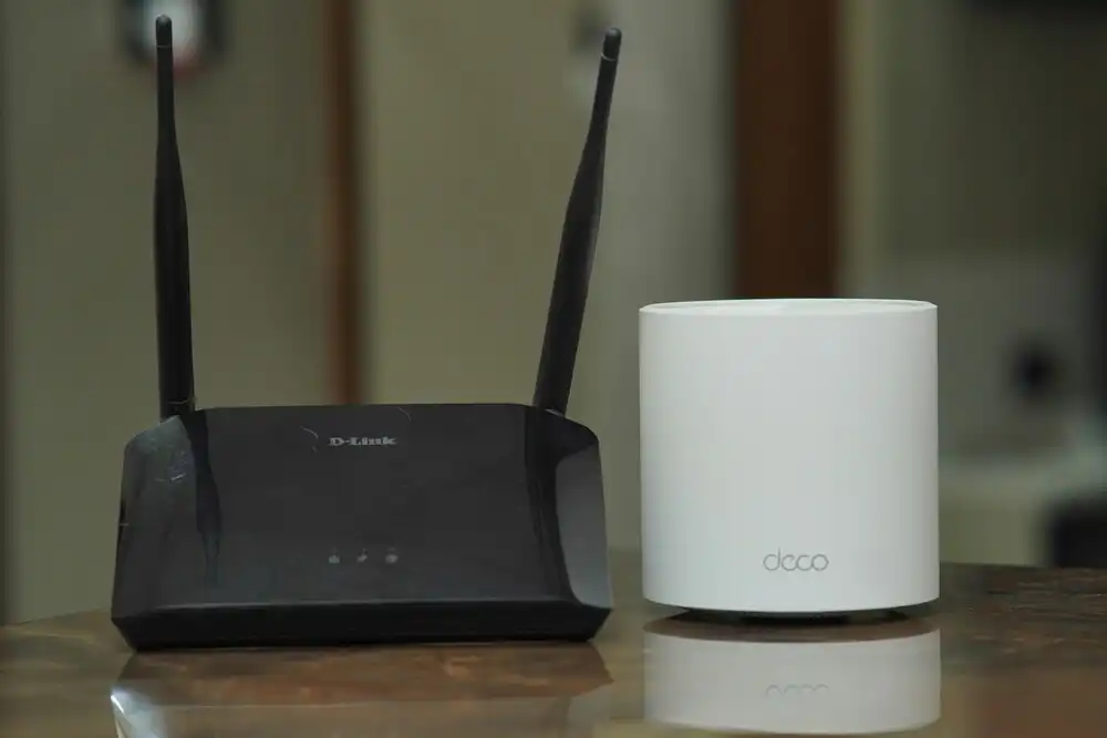 A black D-Link router with two antennas stands next to a cylindrical white Deco device on a wooden surface. The background is blurred, focusing attention on the devices.