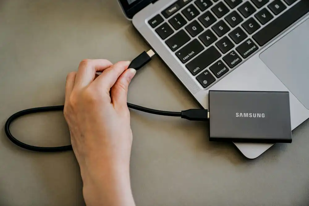 A hand plugging a USB-C cable from a Samsung portable SSD into a silver laptop. The laptop is partially visible on a gray surface.