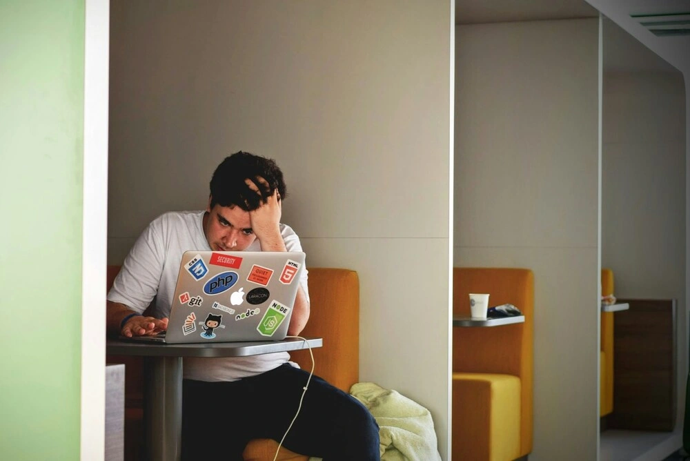 A person sitting at a table in a booth, looking stressed while working on a laptop covered with various tech-related stickers. The setting appears to be indoors with neutral-colored walls and furniture.