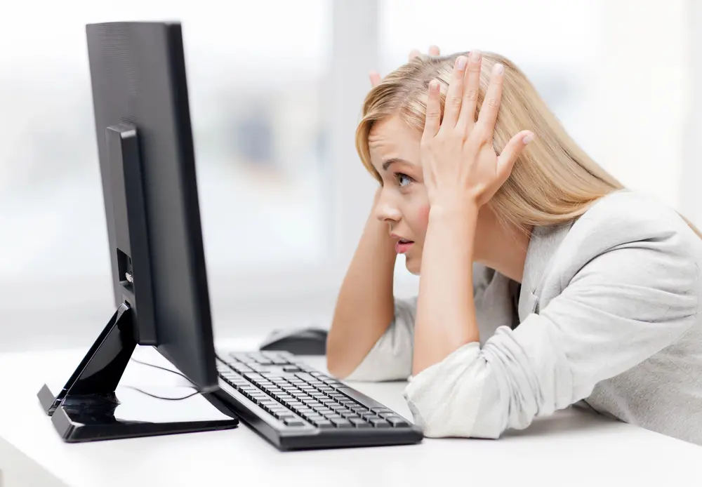 A woman with long blonde hair sitting at a desk looks frustrated, holding her head in her hands while staring at a computer monitor. A keyboard and mouse are placed in front of her, and the background shows a bright, blurred office setting.