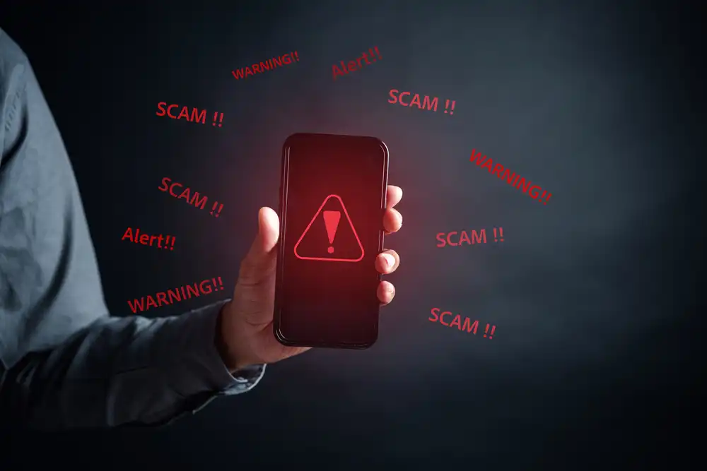 A hand holds a smartphone displaying a red warning symbol with an exclamation mark. The words "SCAM!!", "WARNING!!", and "Alert!!" float around the phone on a dark background, highlighting a digital security threat.