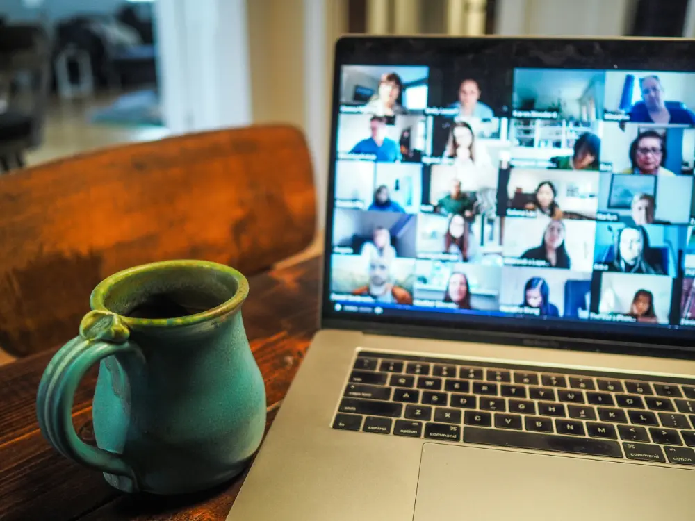 A green ceramic mug sits next to a laptop on a wooden table. The laptop screen displays a virtual meeting with many participants visible in individual video call windows.