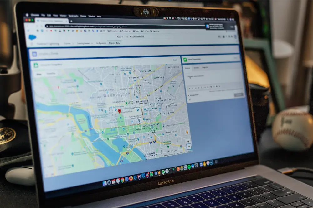A MacBook Pro displays a digital map of Washington, D.C. on its screen, with a location pin marked. The laptop sits on a desk with a baseball and other objects nearby.