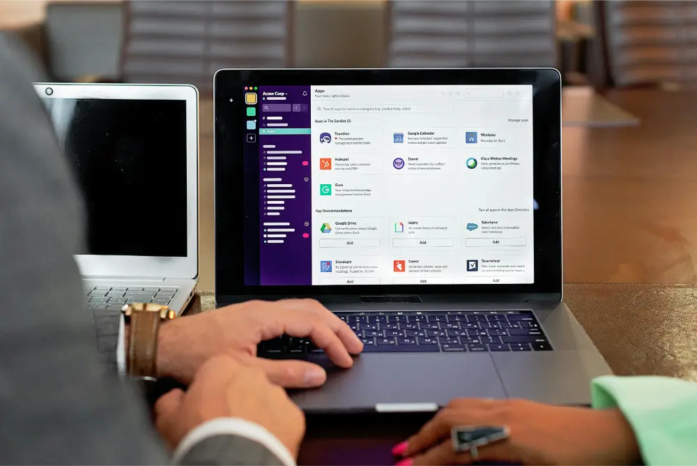 Two people sit at a table using laptops; one screen displays the Slack desktop app with various integrations, and the other laptop is partially visible. Hands are poised on the keyboards, indicating active collaboration.