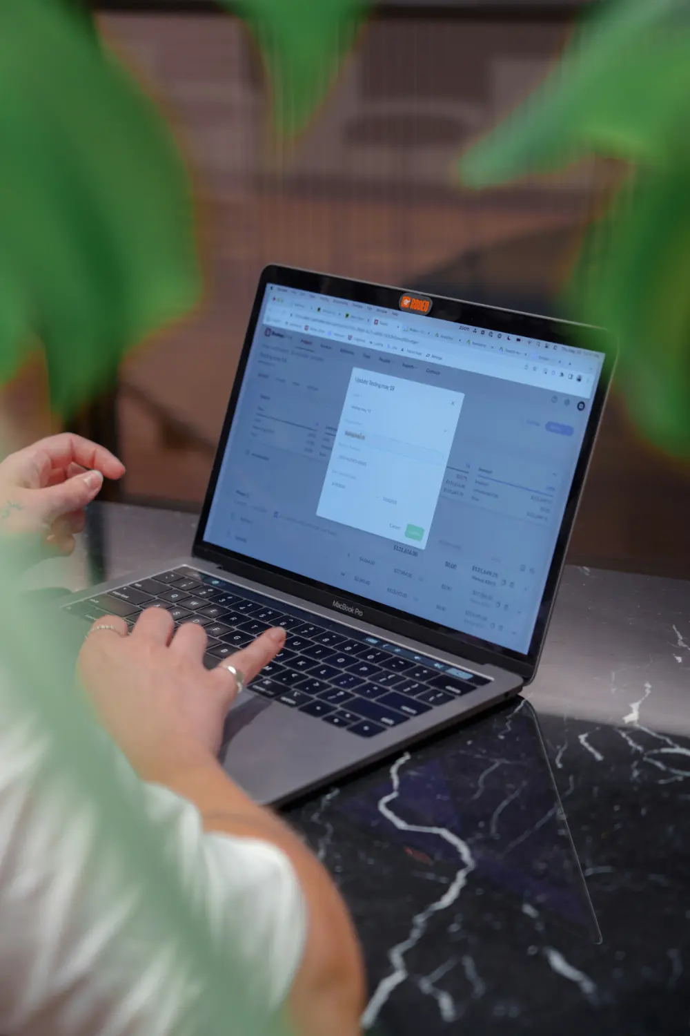 A person uses a laptop on a black marble table, entering information onscreen. Green leaves partially frame the image from the foreground.