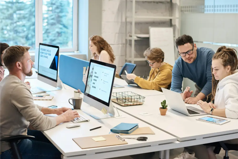 Six people work together in a modern office. Some use desktop computers while others use laptops or tablets. One person stands and talks to a seated colleague. There are office supplies and plants on the desk.