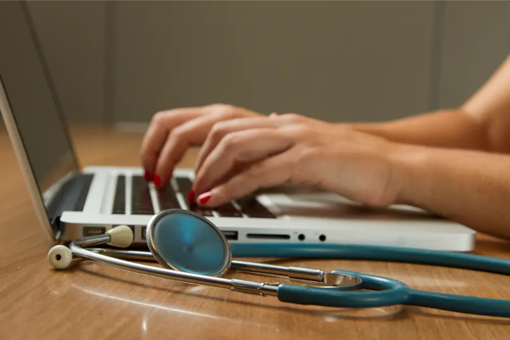 A person with red nail polish types on a laptop at a wooden desk, with a stethoscope placed beside the computer, suggesting a medical or healthcare setting.
