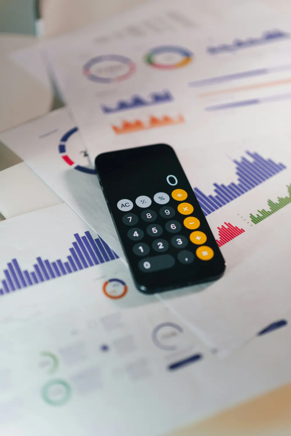 A smartphone with a calculator app open is placed on top of printed sheets displaying colorful bar graphs, charts, and financial data.