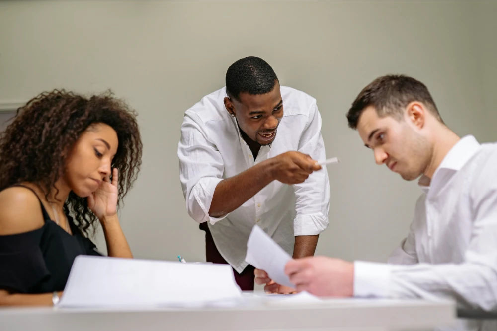 Three colleagues are having a serious discussion at a desk; one man is standing and pointing while the other two, seated, look at documents, with the woman appearing frustrated and the man focused on the paper.