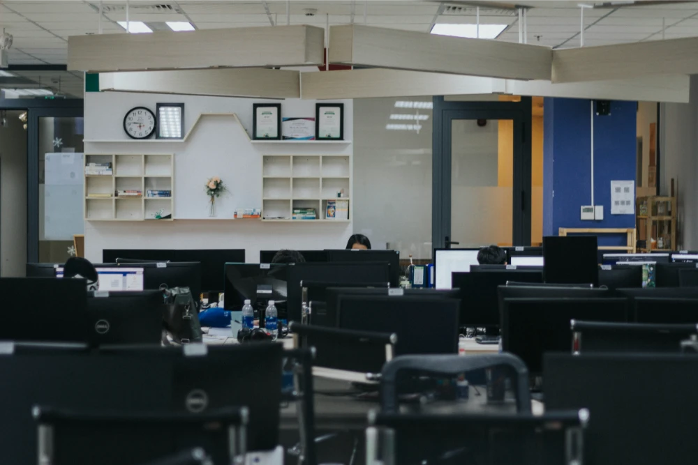 Open office space with several desks and computer monitors; a few people are seated and working. The background features shelves with decorations, framed documents, and a wall clock. The room appears quiet and organized.
