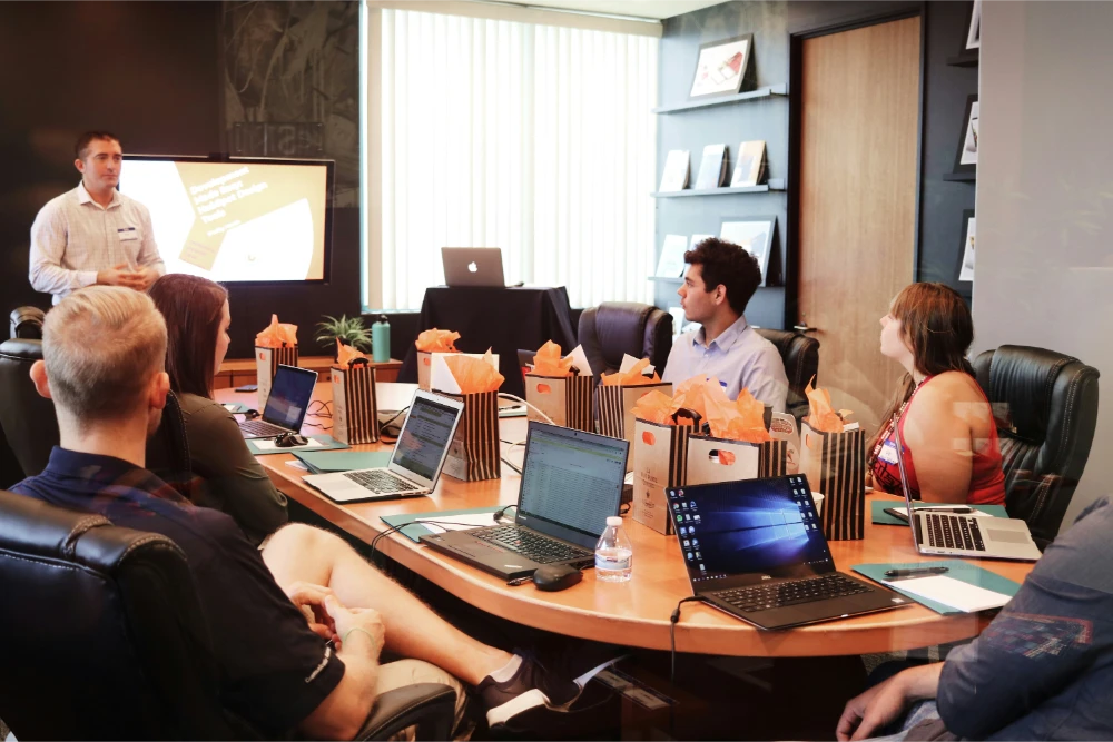 A group of people sit around a conference table with laptops, listening to a presenter standing near a screen displaying a presentation. Gift bags are placed on the table in front of each attendee.