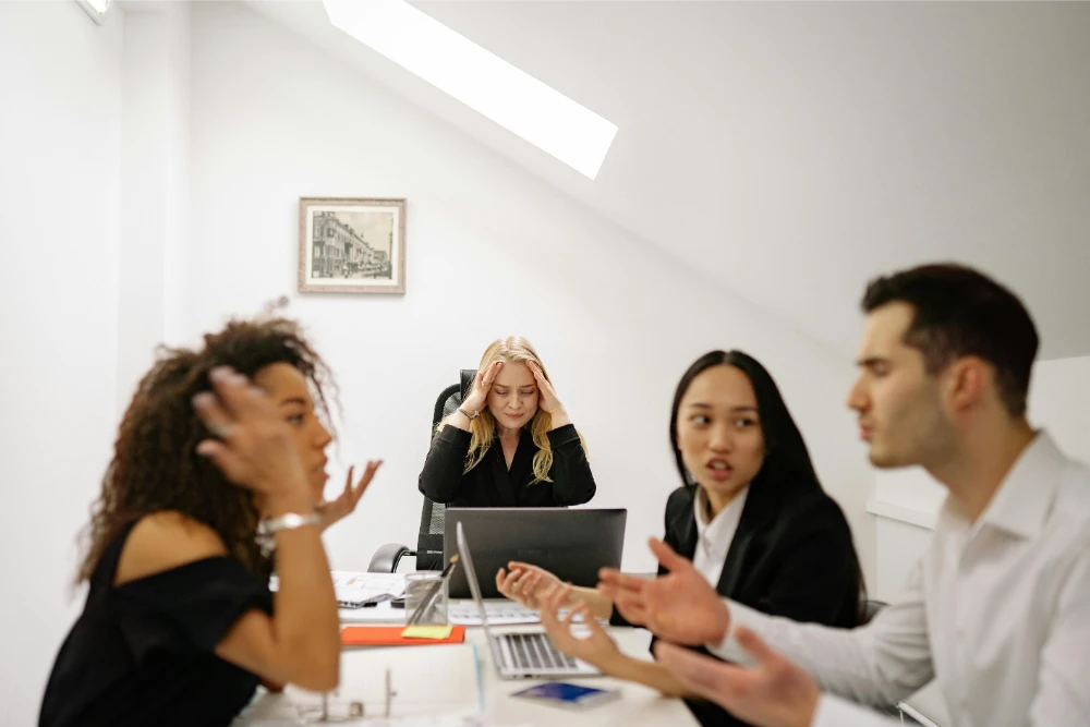 Four colleagues in an office are having a heated discussion, while a woman sitting at the back with a laptop looks stressed, holding her head in her hands.
