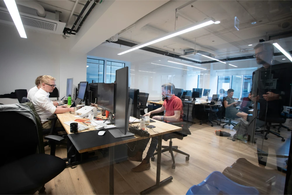 Modern open-plan office with multiple people working at desks equipped with large monitors and laptops. The workspace has wooden floors, white walls, and bright overhead lighting.