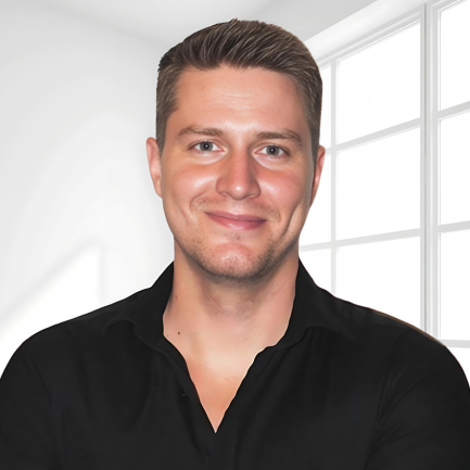 A person with short, neatly styled hair is smiling and looking at the camera. They are wearing a black collared shirt. The background is minimalistic with a white wall and large windows letting in natural light.