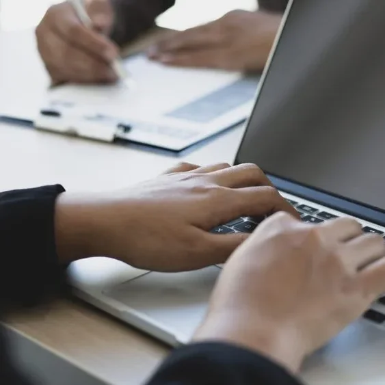 Close-up of hands typing on a laptop keyboard. In the background, a person is writing on a clipboard.