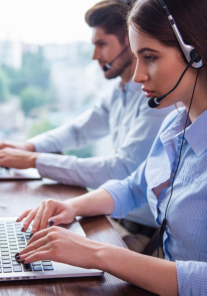 Two customer service representatives are seated at a desk, both wearing headsets and typing on laptops.