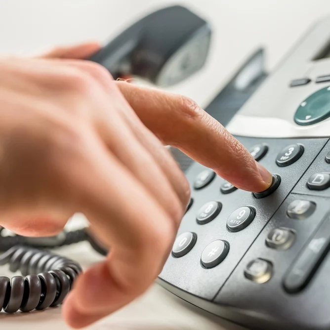 A close-up of a person's hand dialing a number on a landline telephone. The phone is off the hook, and the display and buttons are visible. The finger is pressing a button on the keypad.