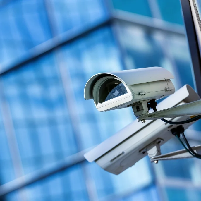 Close-up of a white security camera mounted on a building exterior. The background shows a glass facade reflecting blue and green hues, suggesting an urban setting with modern architecture.