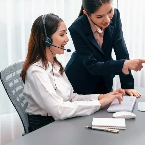 Two women in an office setting, one seated and wearing a headset, typing on a keyboard. The other stands beside her, pointing at the computer screen.