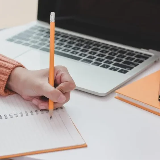 A person in an orange sweater writes in a lined notebook with a pencil. A laptop is open on a round white table.