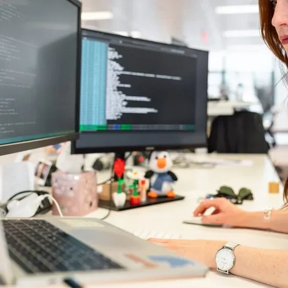 A person with long red hair is working at a white desk with a laptop and two monitors displaying code.