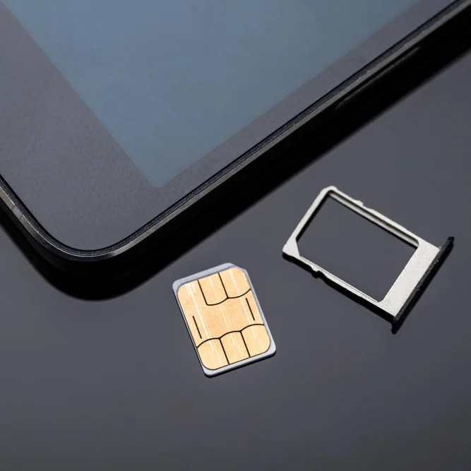 A close-up image of a SIM card and SIM card tray placed on a shiny black surface next to a tablet. The tablet screen is partially visible, with the SIM card and tray positioned beside it.