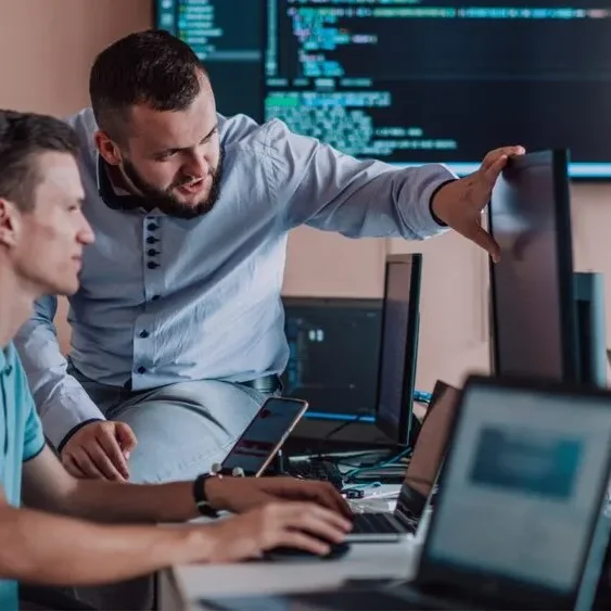 Two men in an office environment work at a desk with multiple computer monitors.