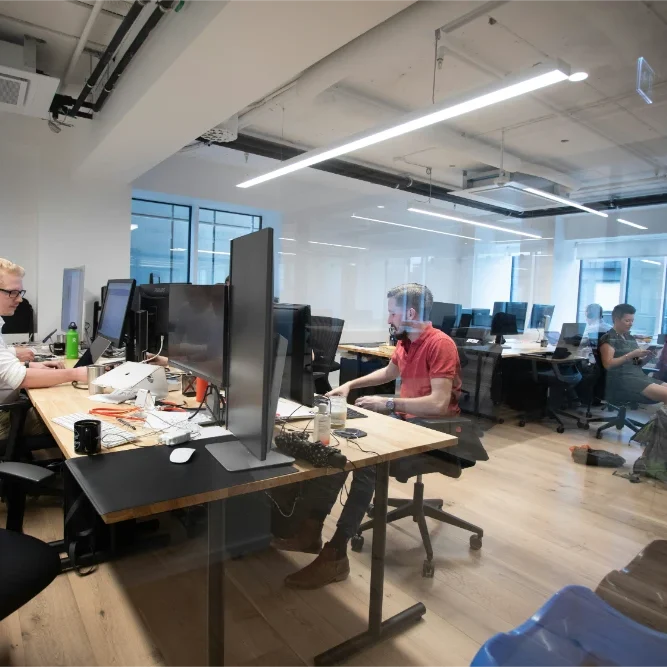 Modern open-plan office with multiple people working at desks equipped with large monitors and laptops. The workspace has wooden floors, white walls, and bright overhead lighting.
