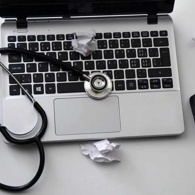 An open laptop with a stethoscope placed on the keyboard, surrounded by crumpled paper balls. A wireless mouse sits to the right of the laptop on a white surface.