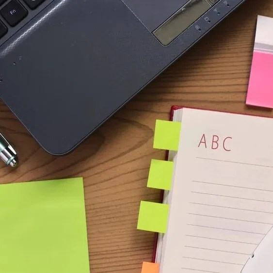 A wooden desk with a laptop, multicolored sticky notes, a pen, and an open notebook with tabbed sections labeled with letters.