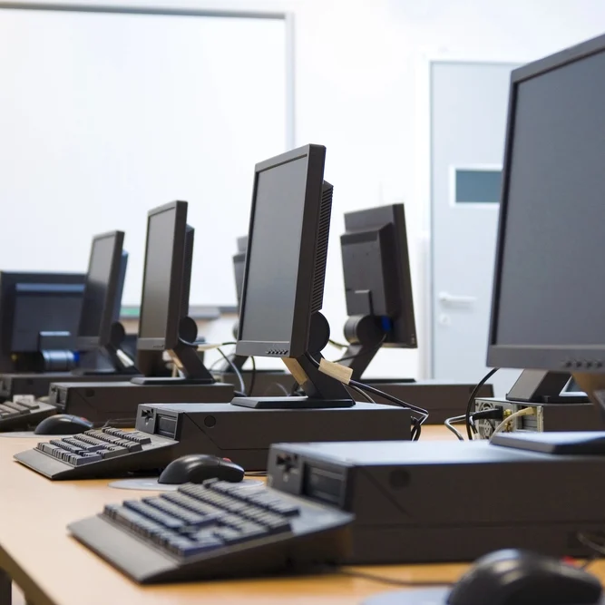 A classroom with several desktop computers placed on wooden tables in a row, each with a monitor, keyboard, and mouse. A whiteboard is visible in the background. The room appears organized and ready for use.