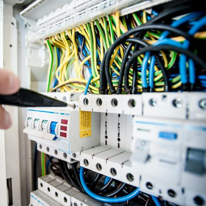 A hand holds a tool near an electrical circuit breaker panel filled with tangled, colorful wires. The circuit breakers and switches are visible, displaying various labels and markings.