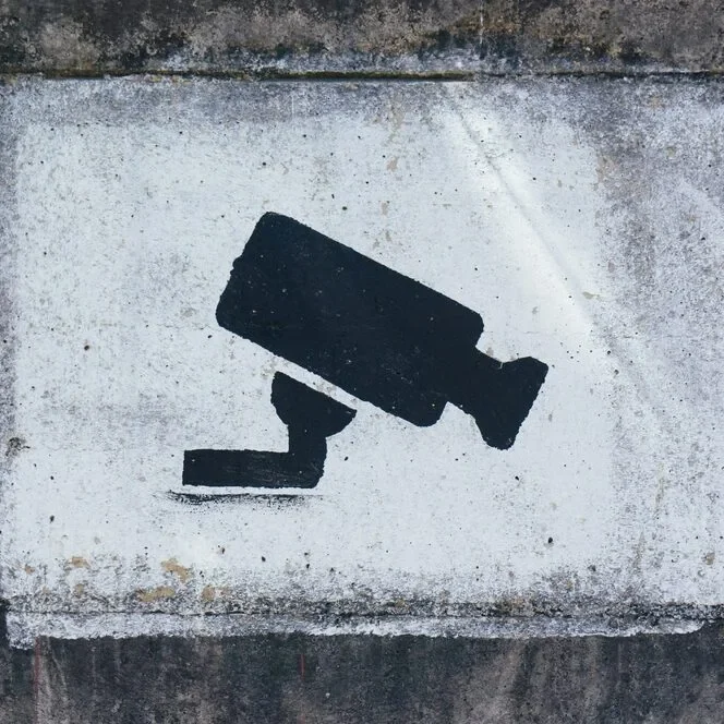 A black silhouette of a surveillance camera is stenciled on a white rectangular background against a rough, dark gray concrete wall.