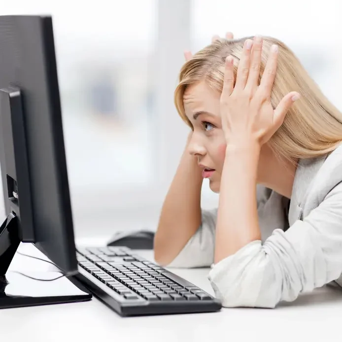 A woman with long blonde hair sitting at a desk looks frustrated, holding her head in her hands while staring at a computer monitor. A keyboard and mouse are placed in front of her, and the background shows a bright, blurred office setting.