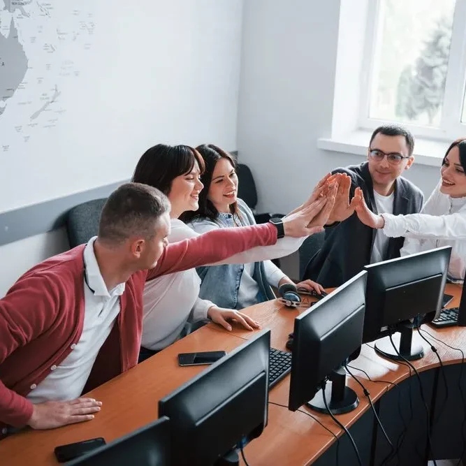 A group of six people, seated at a curved desk with computers, are enthusiastically high-fiving each other.