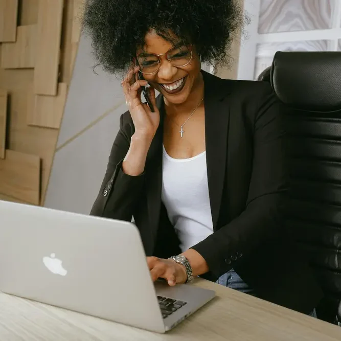 A person with curly hair, wearing glasses and a black blazer, smiles while talking on a cellphone. They are seated at a desk in an office setting, typing on a silver laptop. A coffee cup and saucer are on the desk.