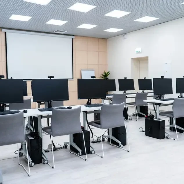 A modern computer lab with multiple workstations arranged in two rows, each equipped with a monitor, keyboard, and mouse. Gray chairs are placed at each station, and a large projector screen is mounted on the front wall. The room is well-lit and organized.