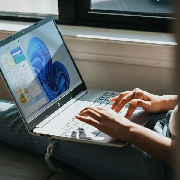 A person is sitting on a windowsill, typing on a silver laptop that displays the Windows 11 operating system on the screen. Sunlight streams through the window, illuminating the scene. The person is wearing casual clothing, including jeans.